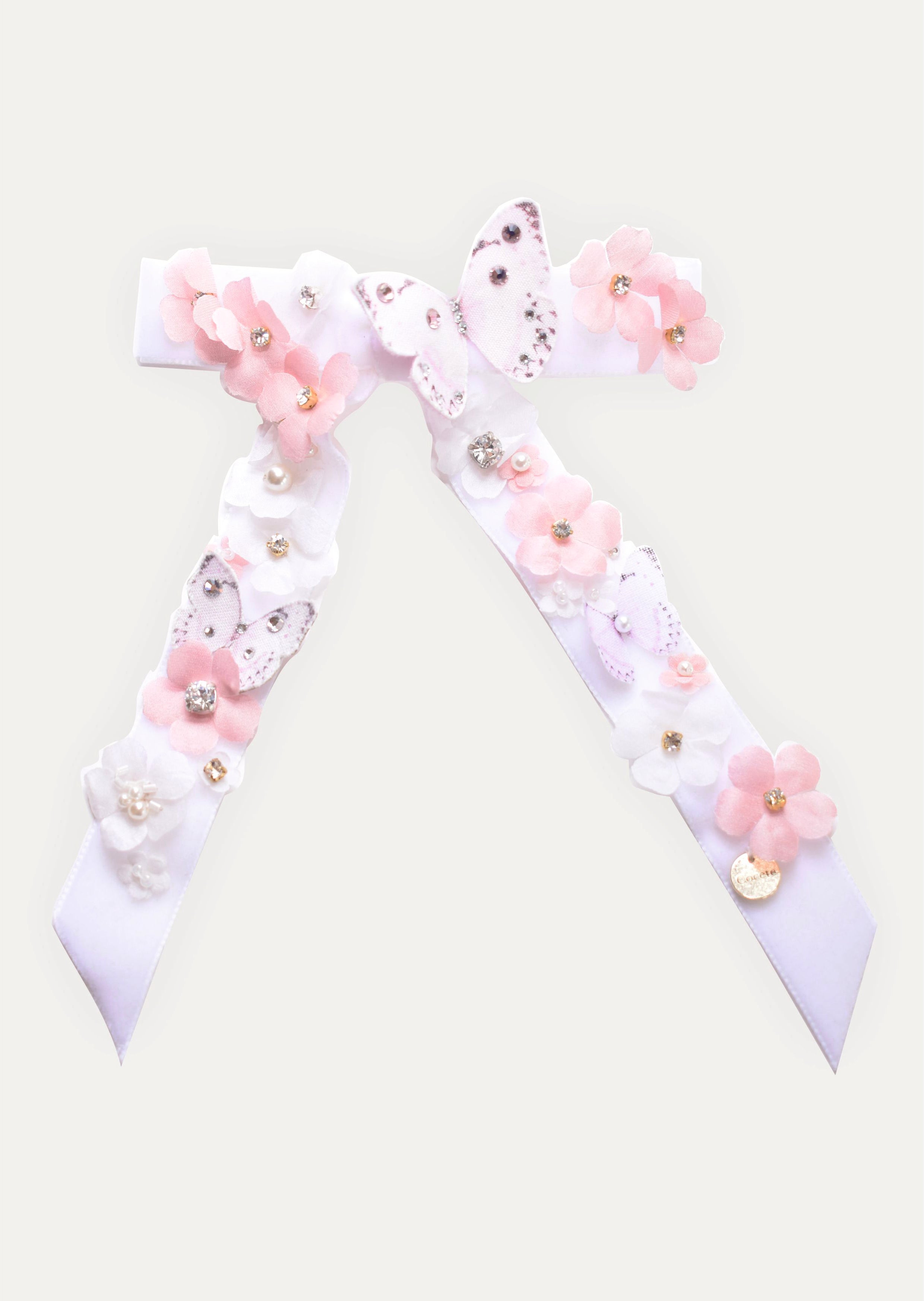 Large handmade white satin hair bow with long tails, decorated with pink flowers, butterflies, pearls, and crystals, featuring a gold COCETE brand charm.
